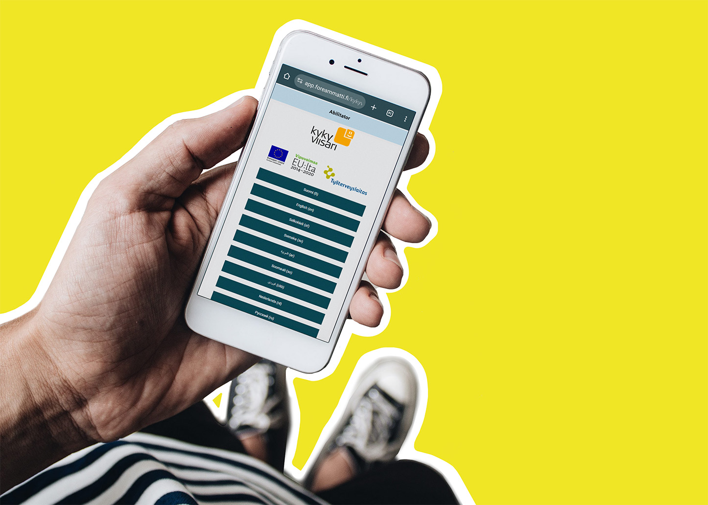 Vasen käsi pitelee älypuhelinta, jonka ruudulla näkyy ForeAmmatin Kykyviisarin kielivalikko. Kuvan tausta on keltainen.